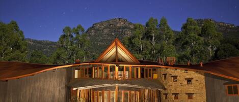 A rustic lodge with a prominent triangular roof, large windows, and a stone facade, set against a mountainous backdrop.
