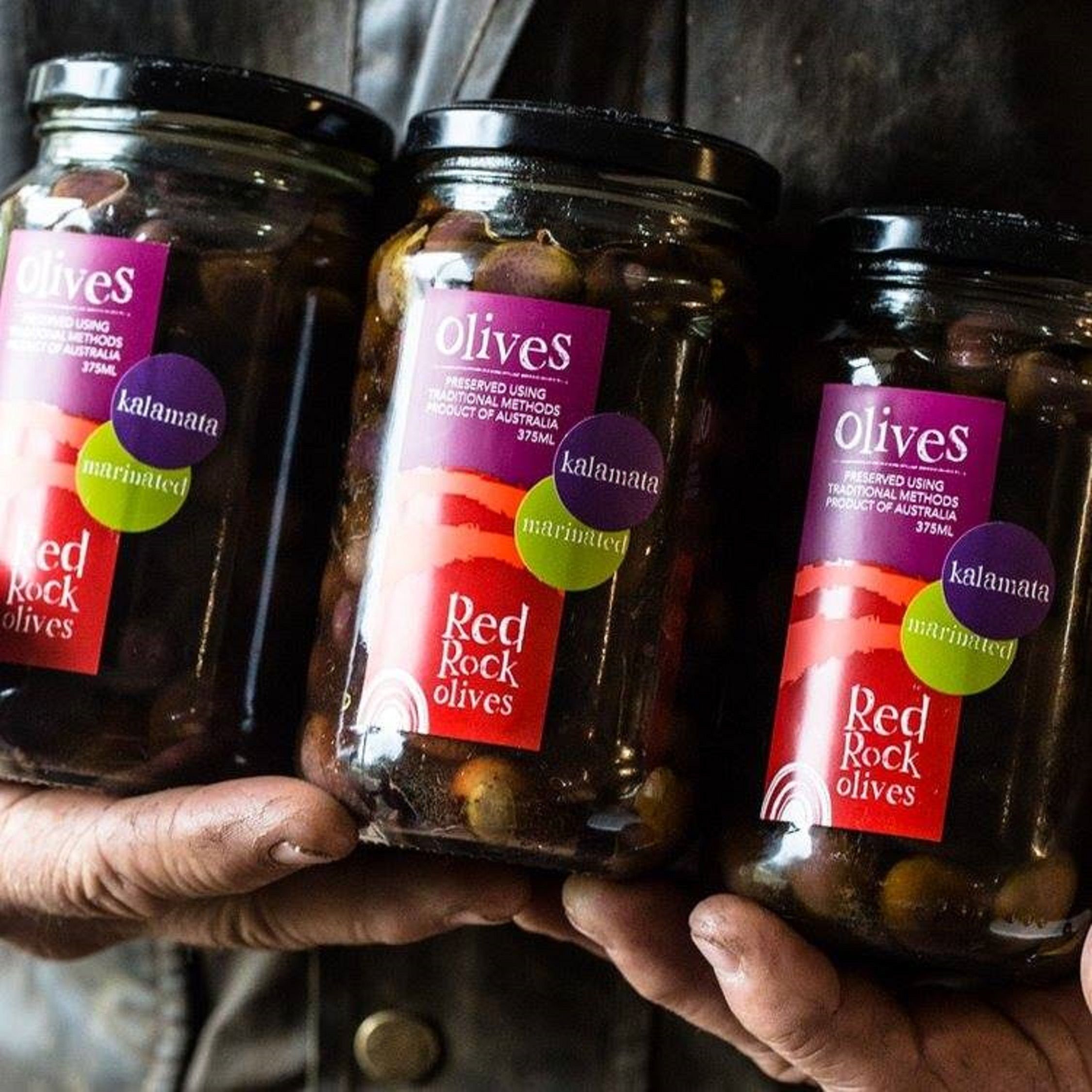 Three jars of Red Rock olives, preserved using traditional methods, held by a person.