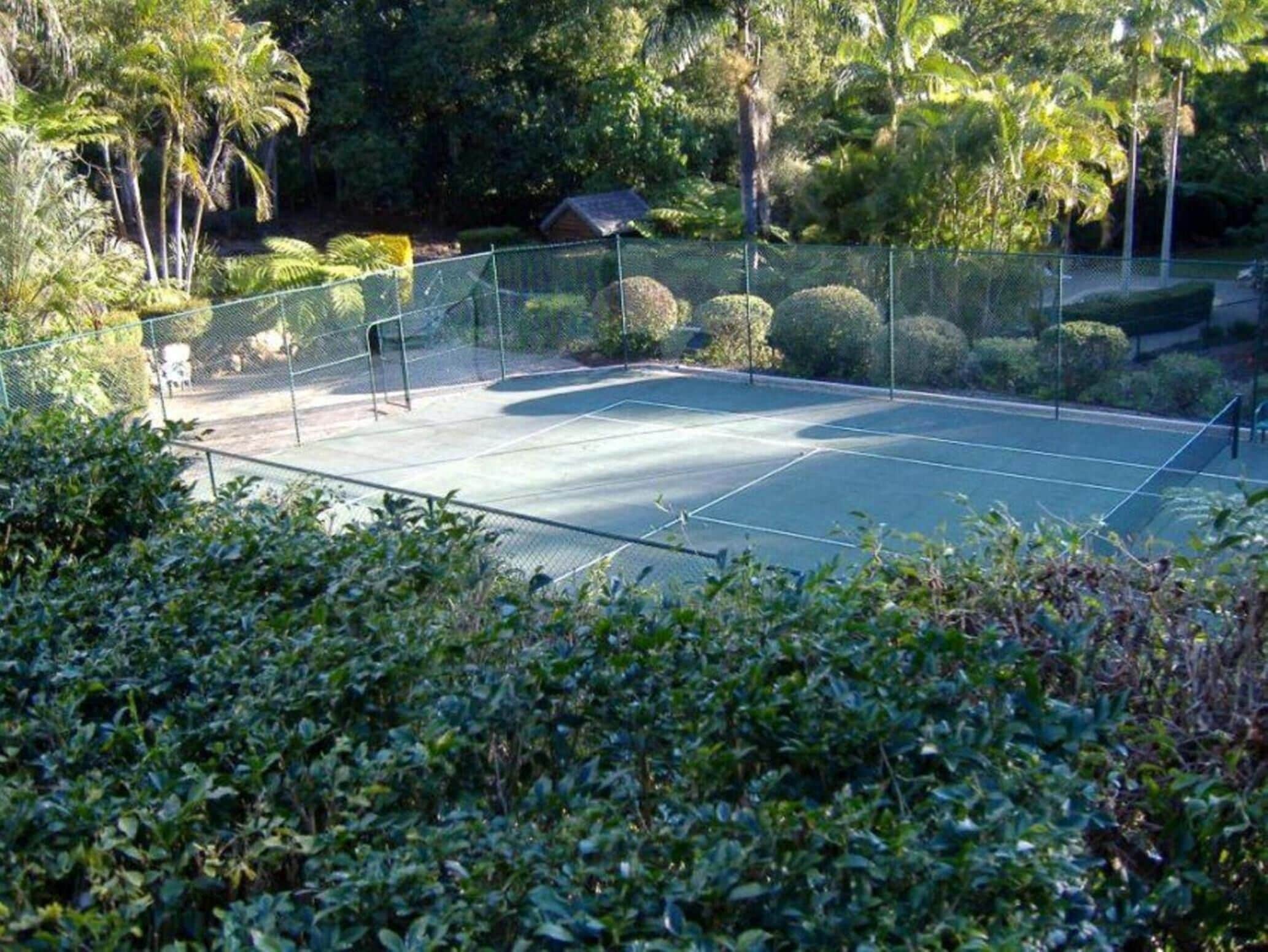 Una cancha de tenis al aire libre rodeada de abundante vegetación y un pequeño edificio al fondo.