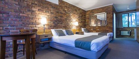 A hotel room with a brick wall, a large bed with a headboard, two bedside tables with lamps, a small table with a chair, and a large window with a view of trees.