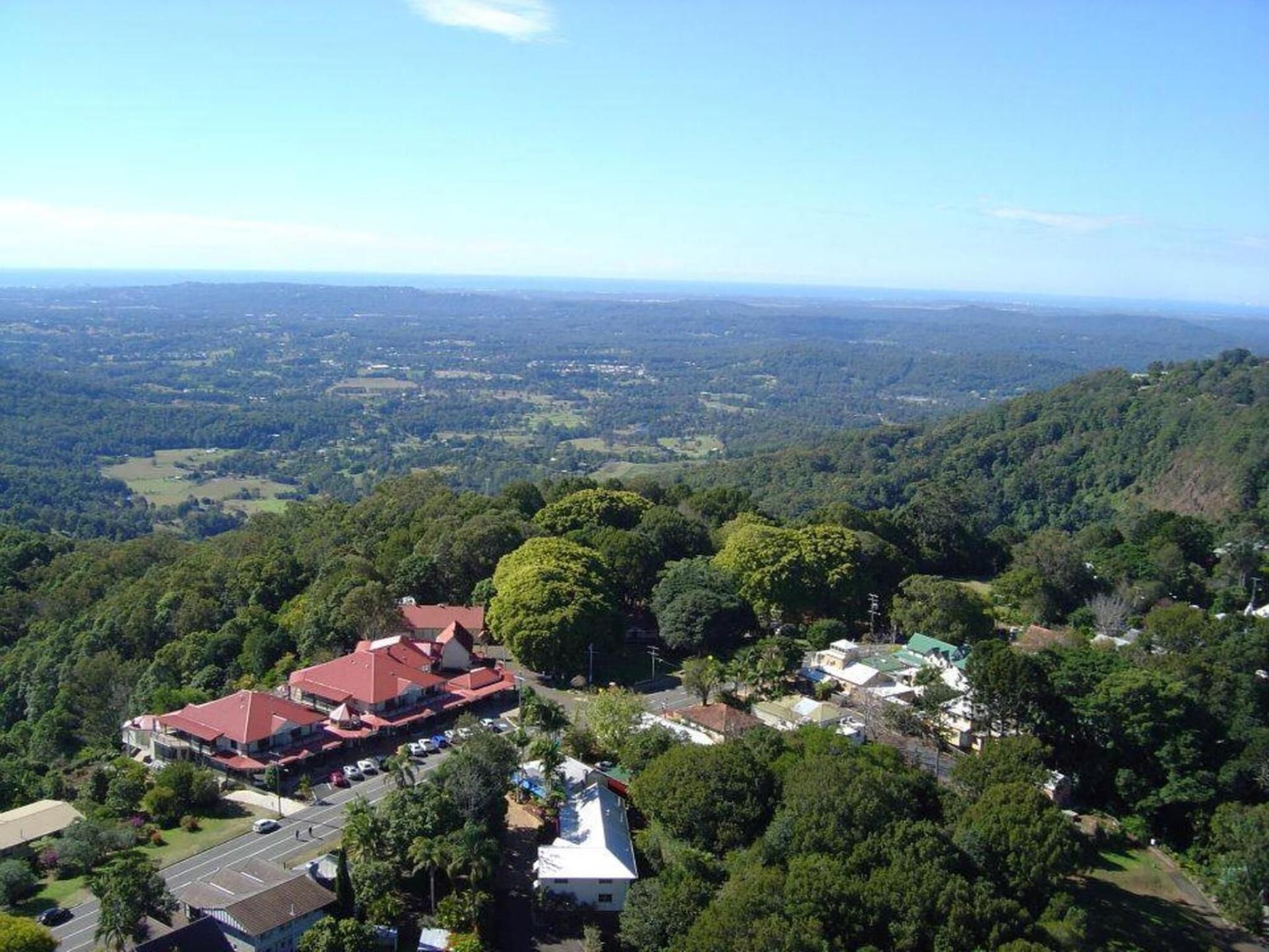 An aerial view of a residential area with houses surrounded by dense forest, a main road, and a clear sky.