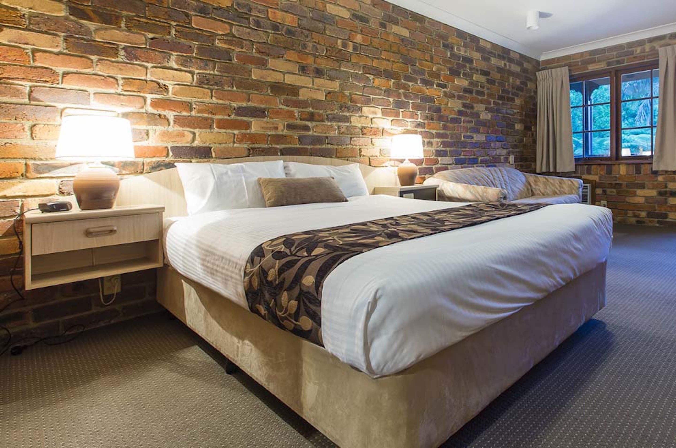 Un dormitorio con pared de ladrillo, una cama grande con colcha estampada, dos lámparas de noche y una ventana con cortinas.