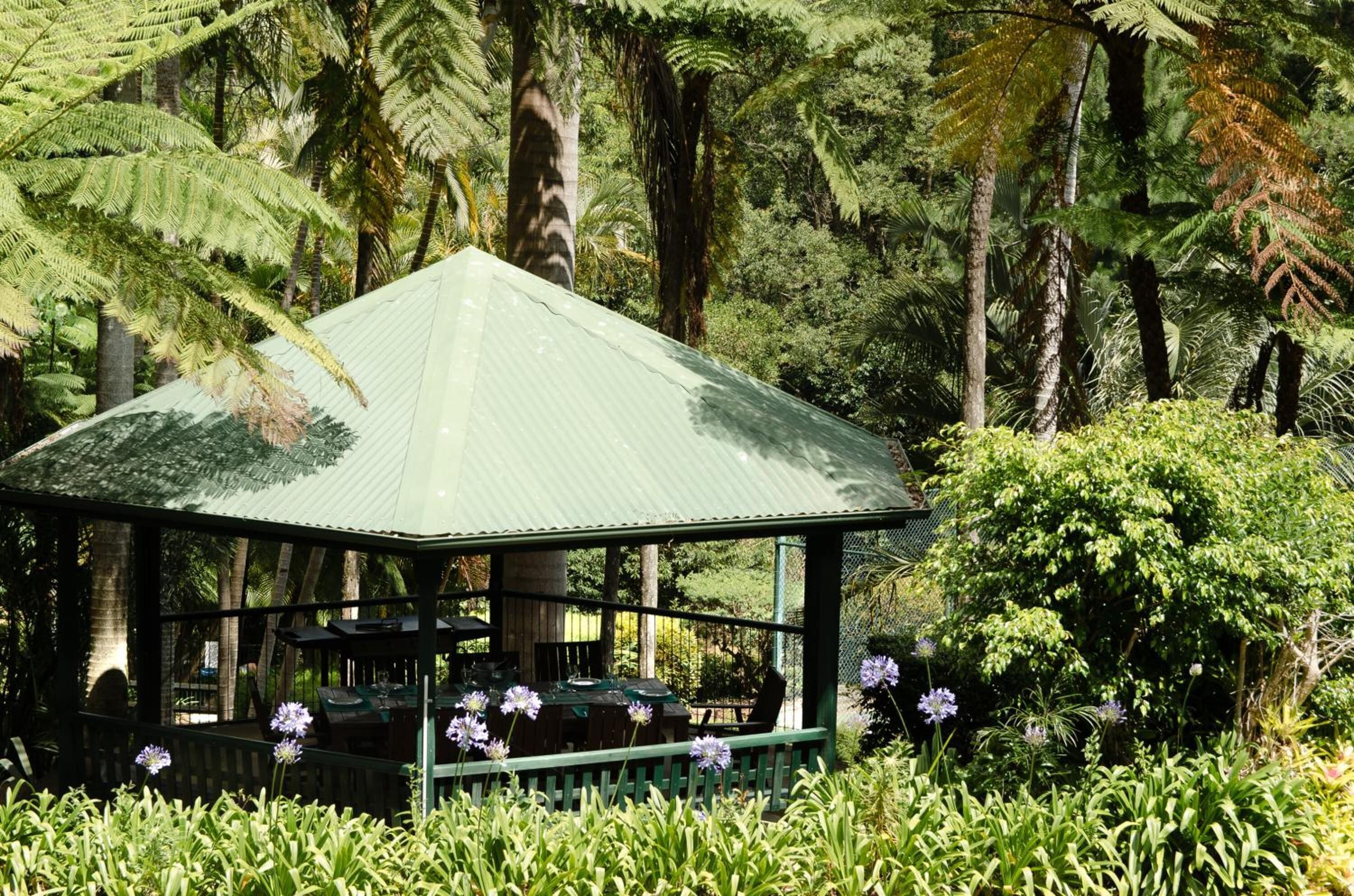 A gazebo with a metal roof surrounded by lush greenery and flowering plants.