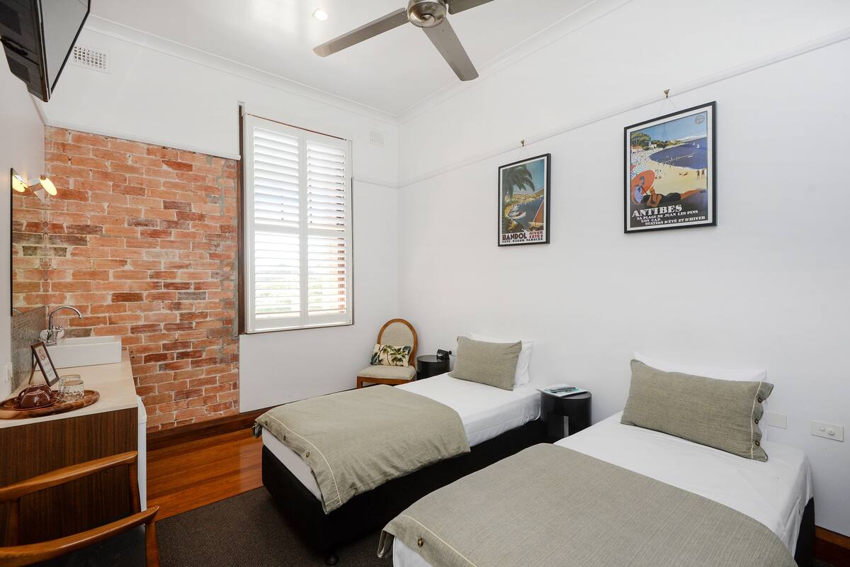 A hotel room with two beds, a brick wall, a window with blinds, a desk with a chair, and a ceiling fan.