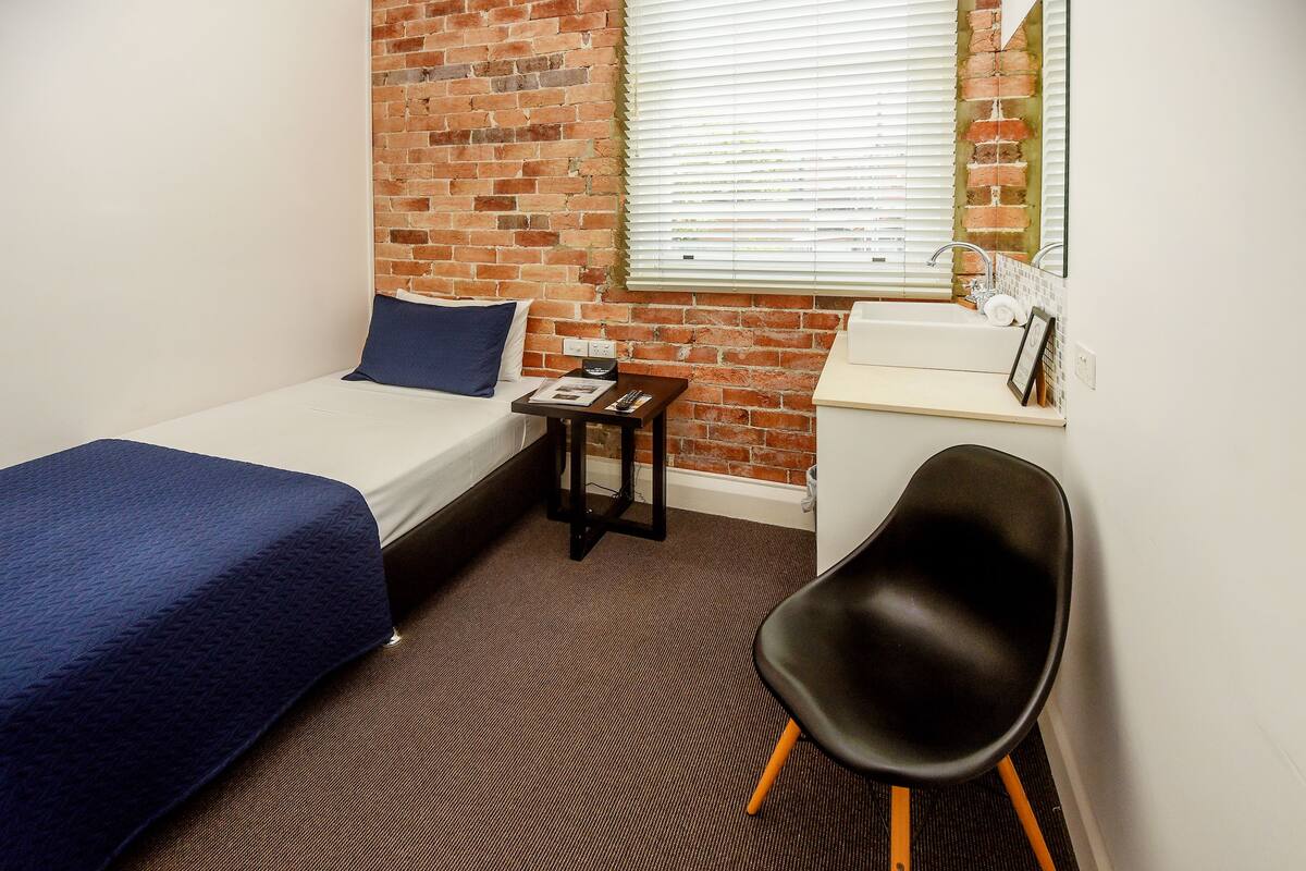 A small, neatly arranged room with a brick wall, a single bed with a blue bedspread, a small table with a book and a remote, a black chair, and a window with blinds.