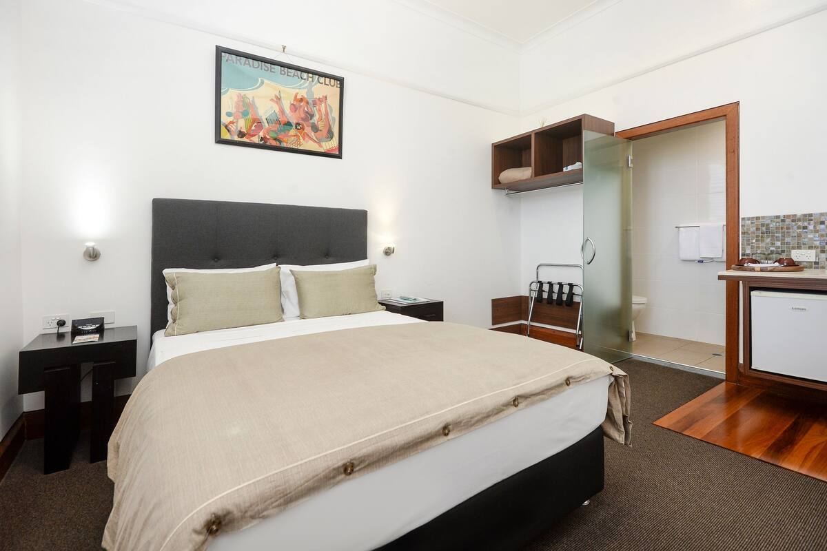 A hotel room with a bed, bedside table, nightstand, and a bathroom visible through a glass door.
