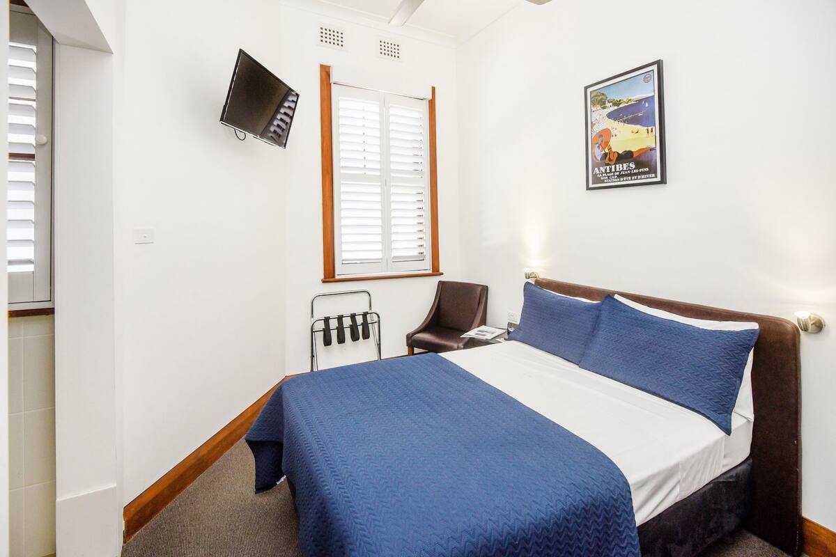 A hotel room with a bed, a chair, a TV, a window with blinds, and a poster on the wall.