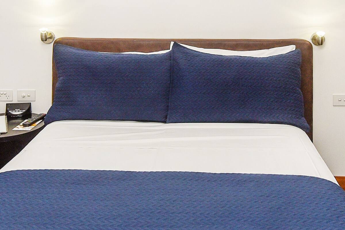 A bed with a white mattress and blue bedding, a nightstand with a phone and remote control, and a wall socket.