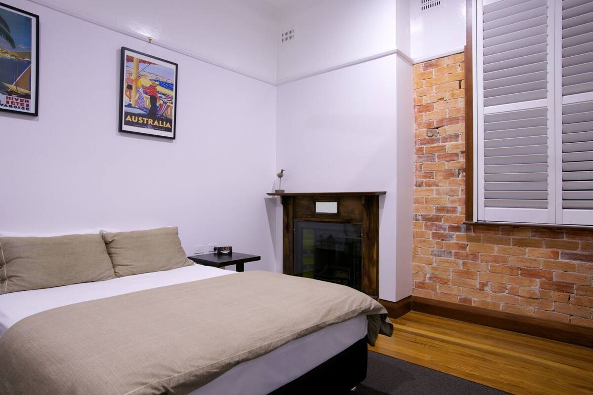 A bedroom with a bed, a small table with a clock, a fireplace, and a brick wall.