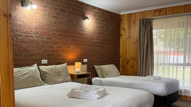 A hotel room with two beds, a brick wall, wooden paneling, a window with curtains, and a nightstand with a lamp. - Country Roads Motor Inn Narrandera (Narrandera)