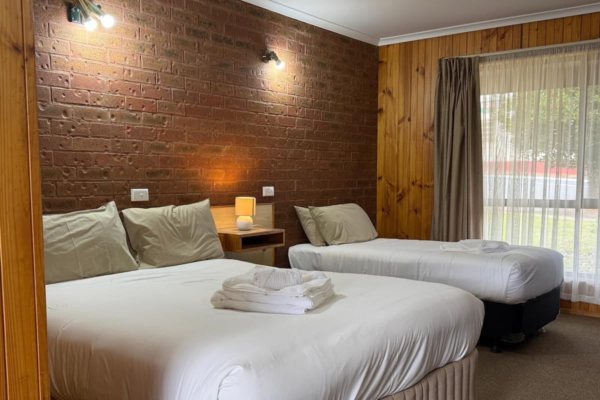 Habitació d'hotel amb dos llits, una paret de maó, revestiment de fusta, una finestra amb cortines i una tauleta de nit amb una làmpada.