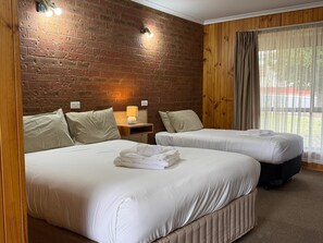 A hotel room with two beds, a brick wall, wooden paneling, a window with curtains, and a nightstand with a lamp. - Country Roads Motor Inn Narrandera (Narrandera)