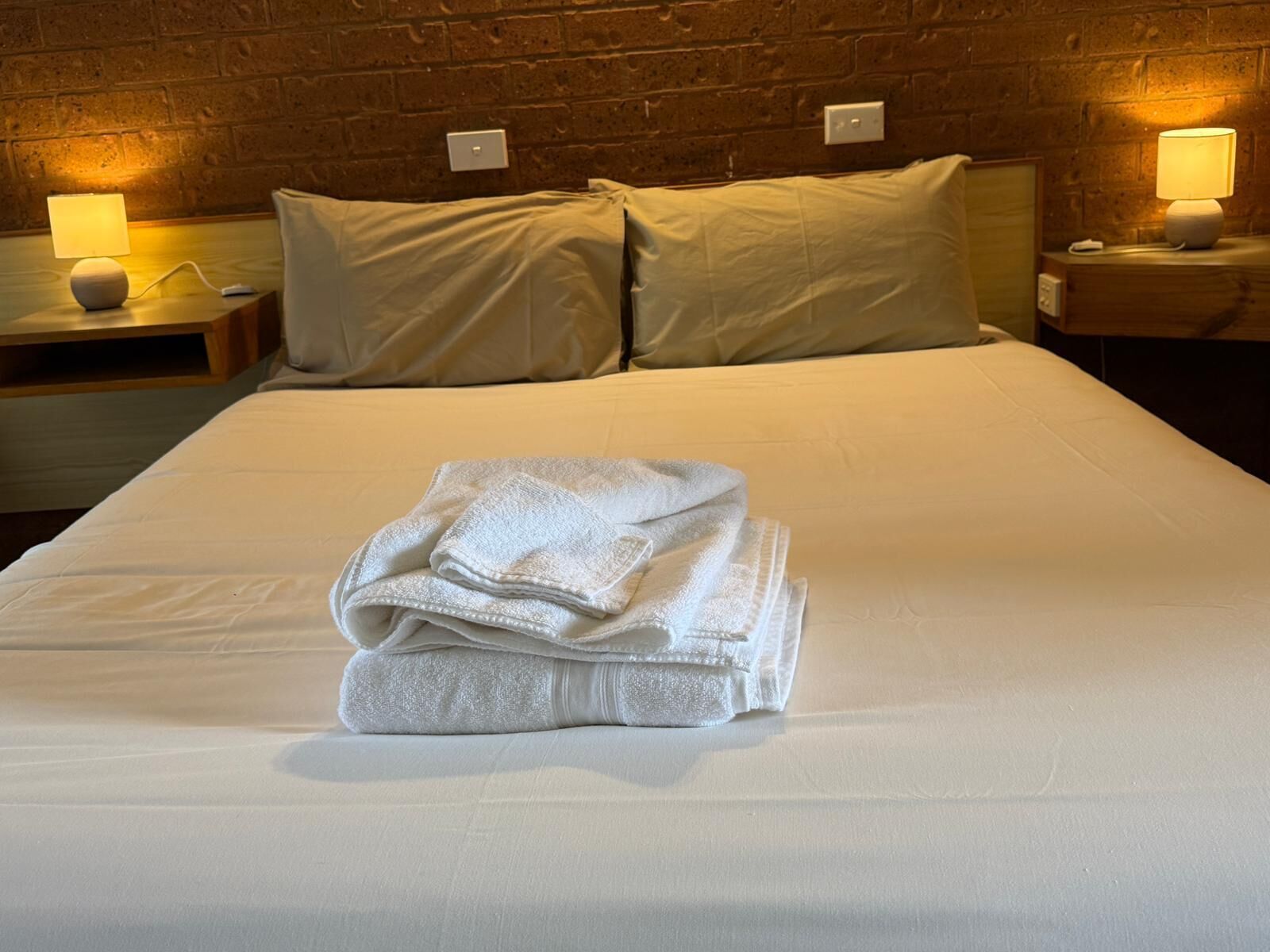 Una cama bien hecha con toallas dobladas, dos lámparas de noche y una pared de ladrillo al fondo.