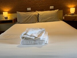 Una cama bien hecha con toallas dobladas, dos lámparas de noche y una pared de ladrillo al fondo.