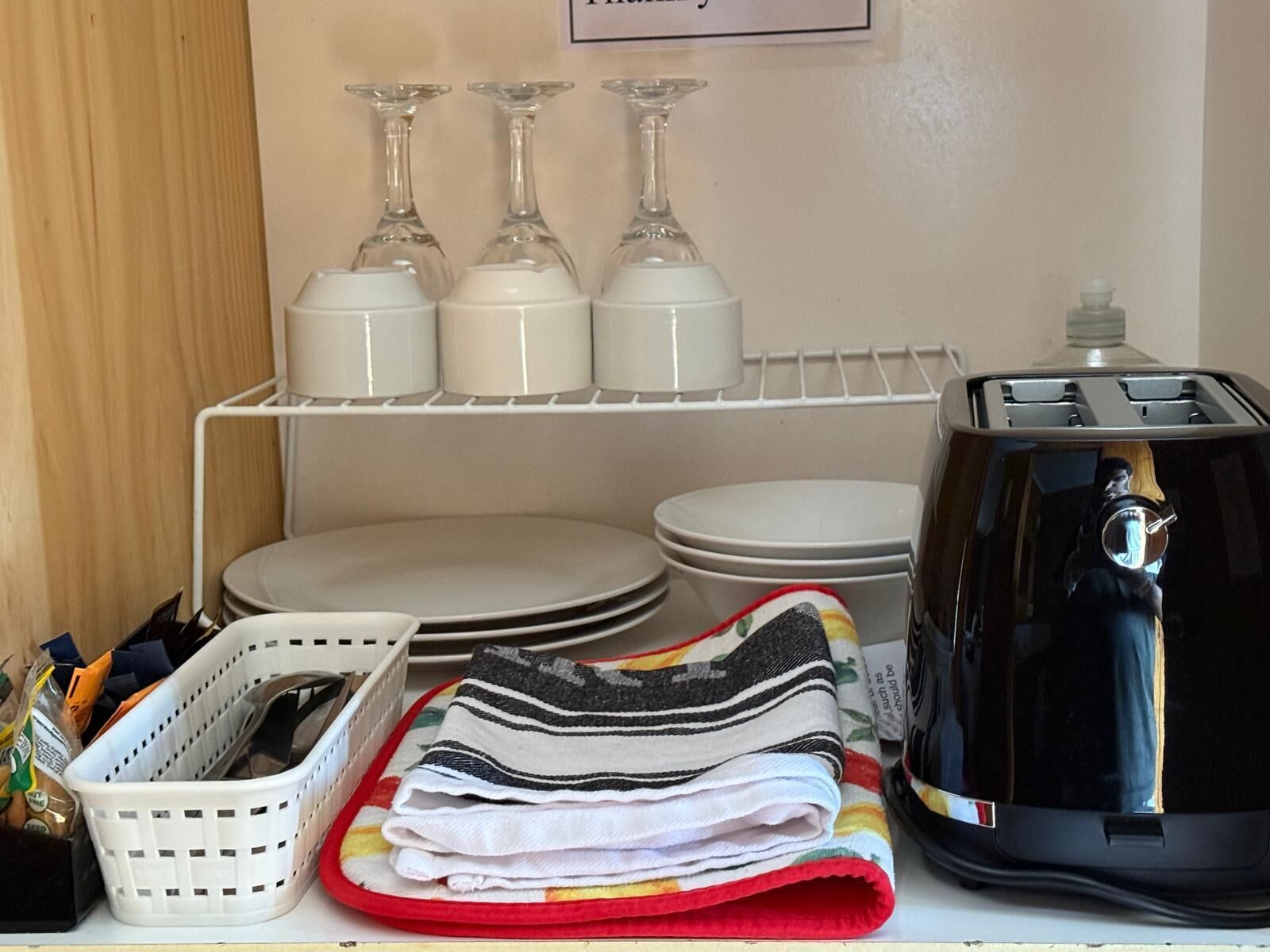 Una zona de cocina con un estante blanco que sostiene copas de vino, platos y un tostador. Hay una cesta con provisiones y toallas dobladas en la encimera.