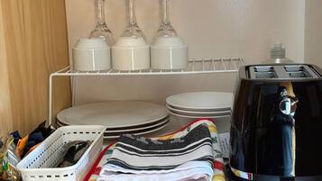 Una zona de cocina con un estante blanco que sostiene copas de vino, platos y un tostador. Hay una cesta con provisiones y toallas dobladas en la encimera.
