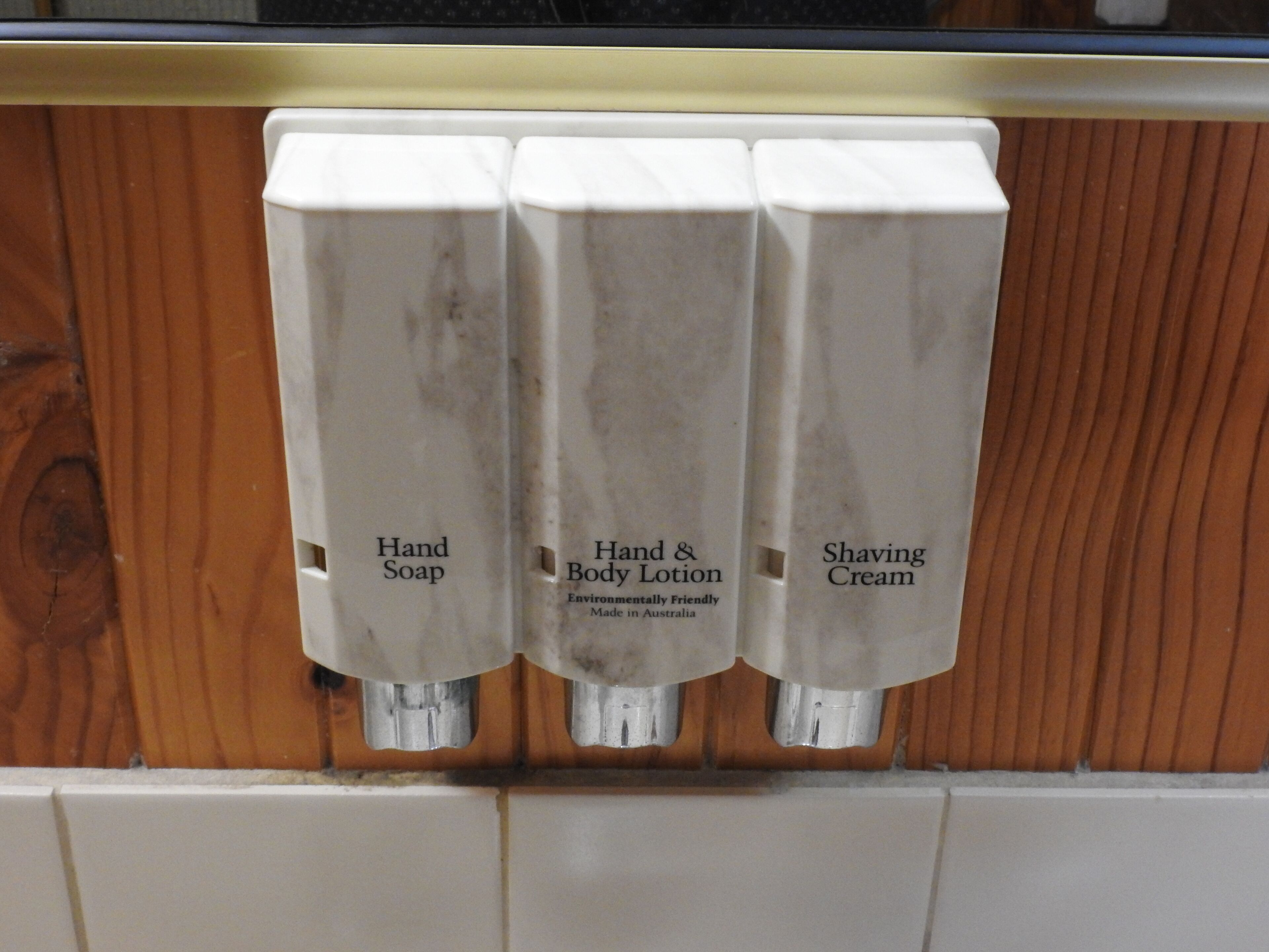 Un distributeur de savon pour les mains, un distributeur de lotion pour les mains et le corps, et un distributeur de mousse à raser, le tout fixé sur une surface en bois.