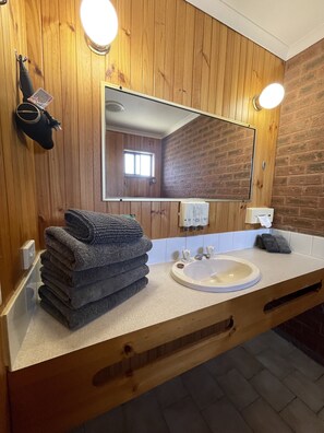 A bathroom with a wooden vanity, a sink, and a mirror. There are towels stacked on the vanity, and a hand dryer is mounted on the wall.