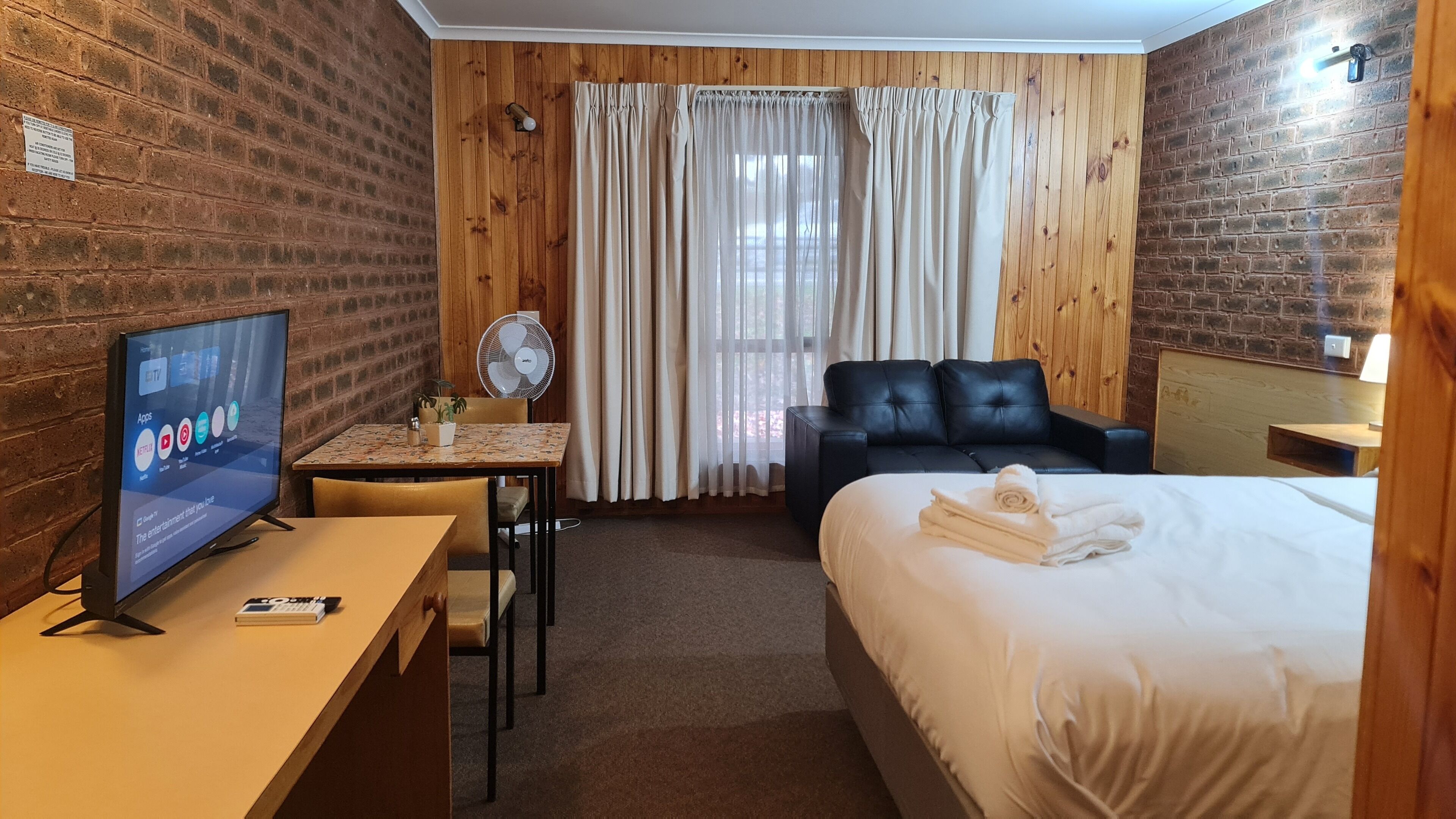 Habitación de hotel con pared de ladrillo, panel de madera, televisor, escritorio con silla, cama con cabecera de cuero negro y ventana con cortinas.