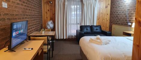 Habitación de hotel con pared de ladrillo, panel de madera, televisor, escritorio con silla, cama con cabecera de cuero negro y ventana con cortinas.