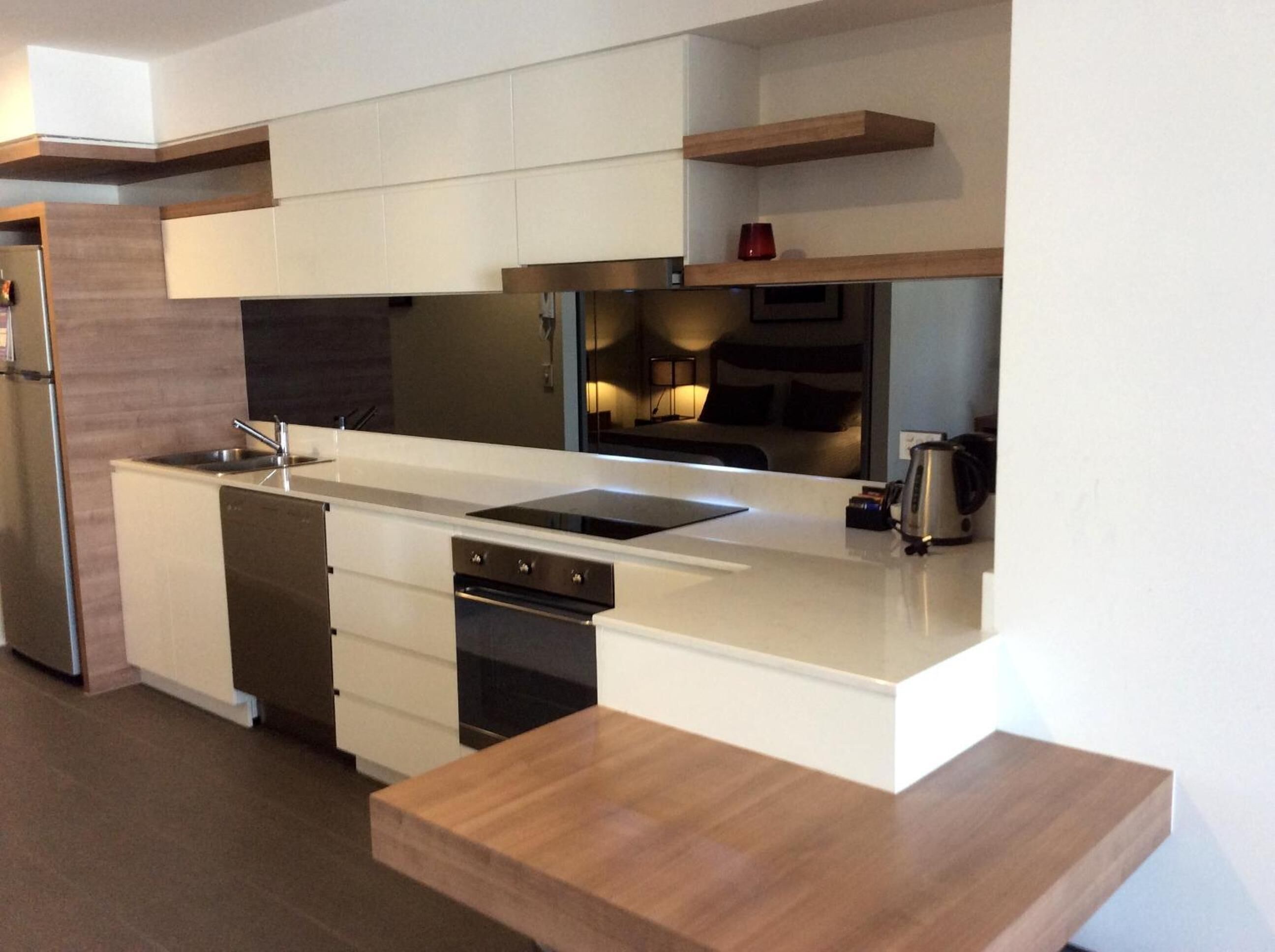 A modern kitchen with white cabinetry, a stainless steel sink, and a built-in oven. There are wooden shelves above the counter and a coffee maker on the right.
