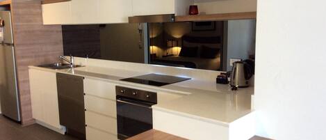 A modern kitchen with white cabinetry, a stainless steel sink, and a built-in oven. There are wooden shelves above the counter and a coffee maker on the right.
