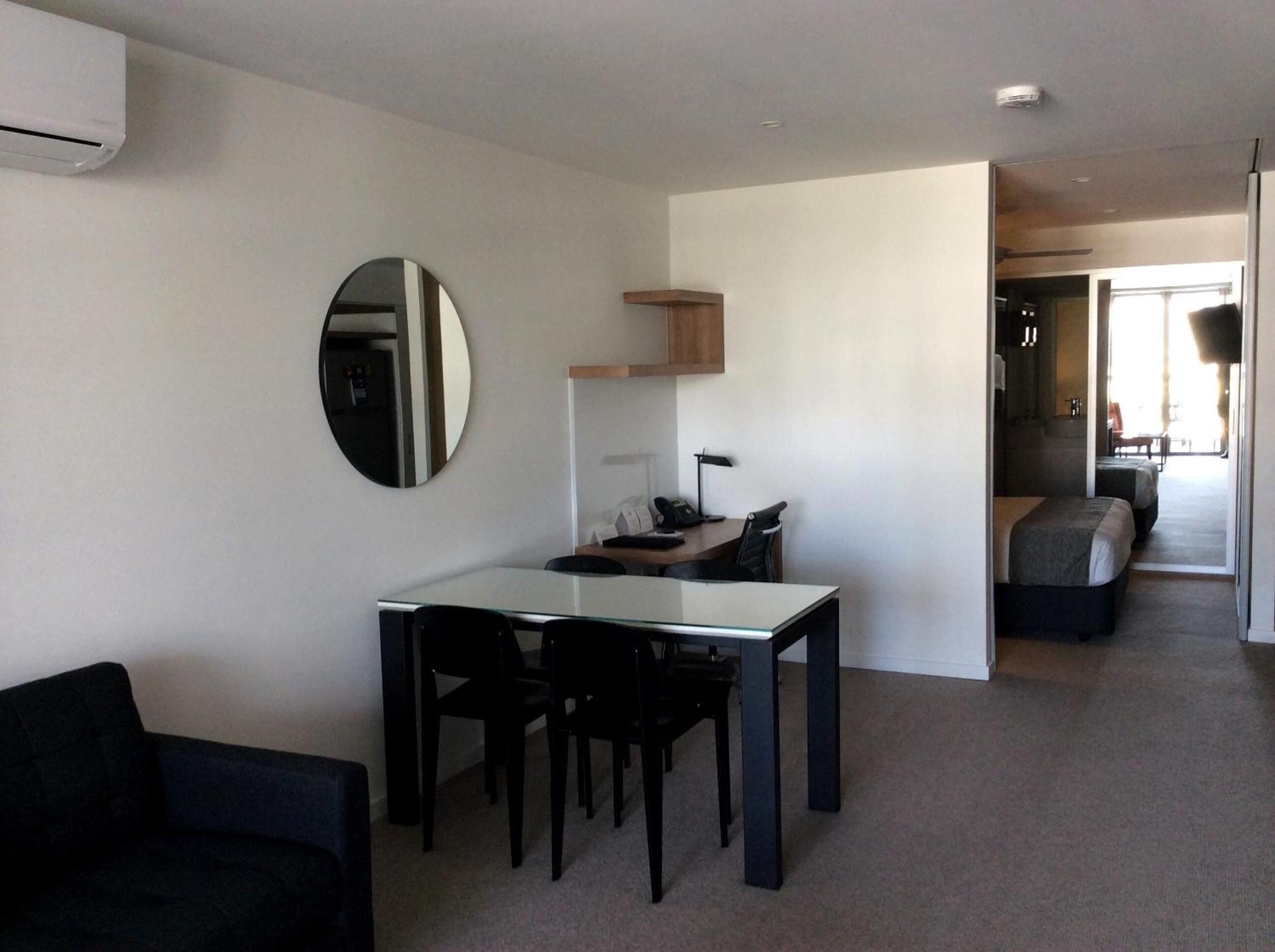 A modern, well-lit room with a dining table and chairs, a small kitchenette, and a bed in the background.