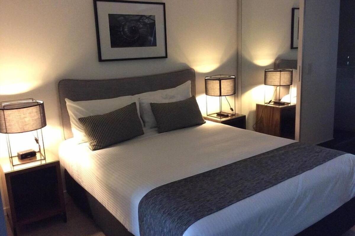 A neatly made bed with a headboard, two bedside tables with lamps, a framed picture on the wall, and a mirror on the wall.