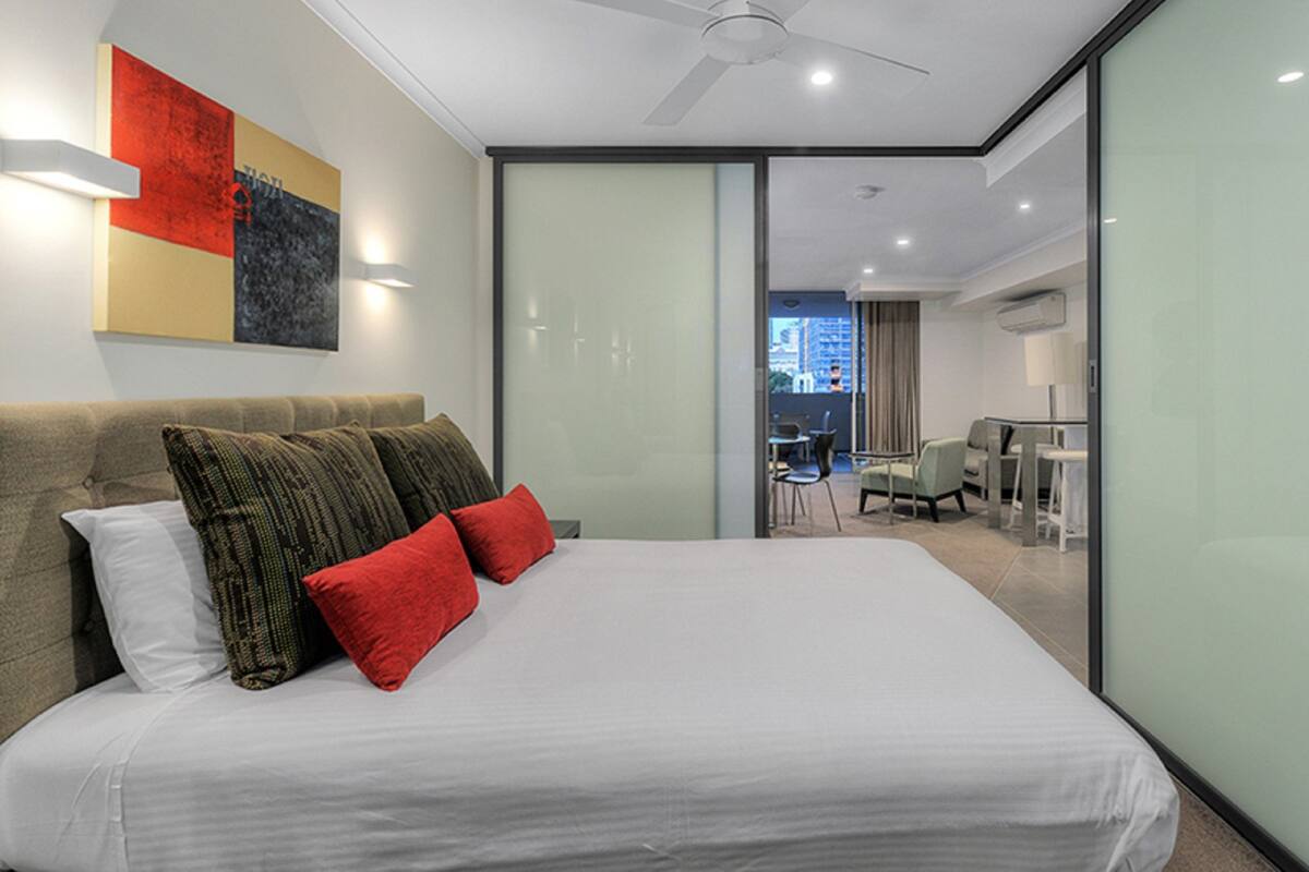 A modern hotel room with a large bed, a ceiling fan, and a city view through a glass wall.