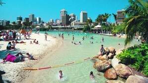 Une plage à l’eau claire, où l’on peut voir des gens nager et bronzer, avec une vue sur les bâtiments d’une ville en arrière-plan.