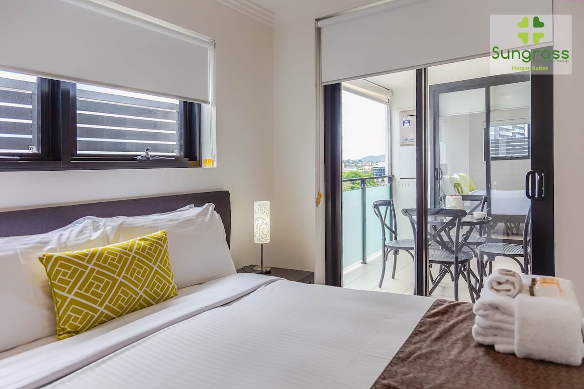 A hotel room with a bed, a nightstand, a lamp, a window with blinds, and a view of an outdoor balcony with chairs and a table.