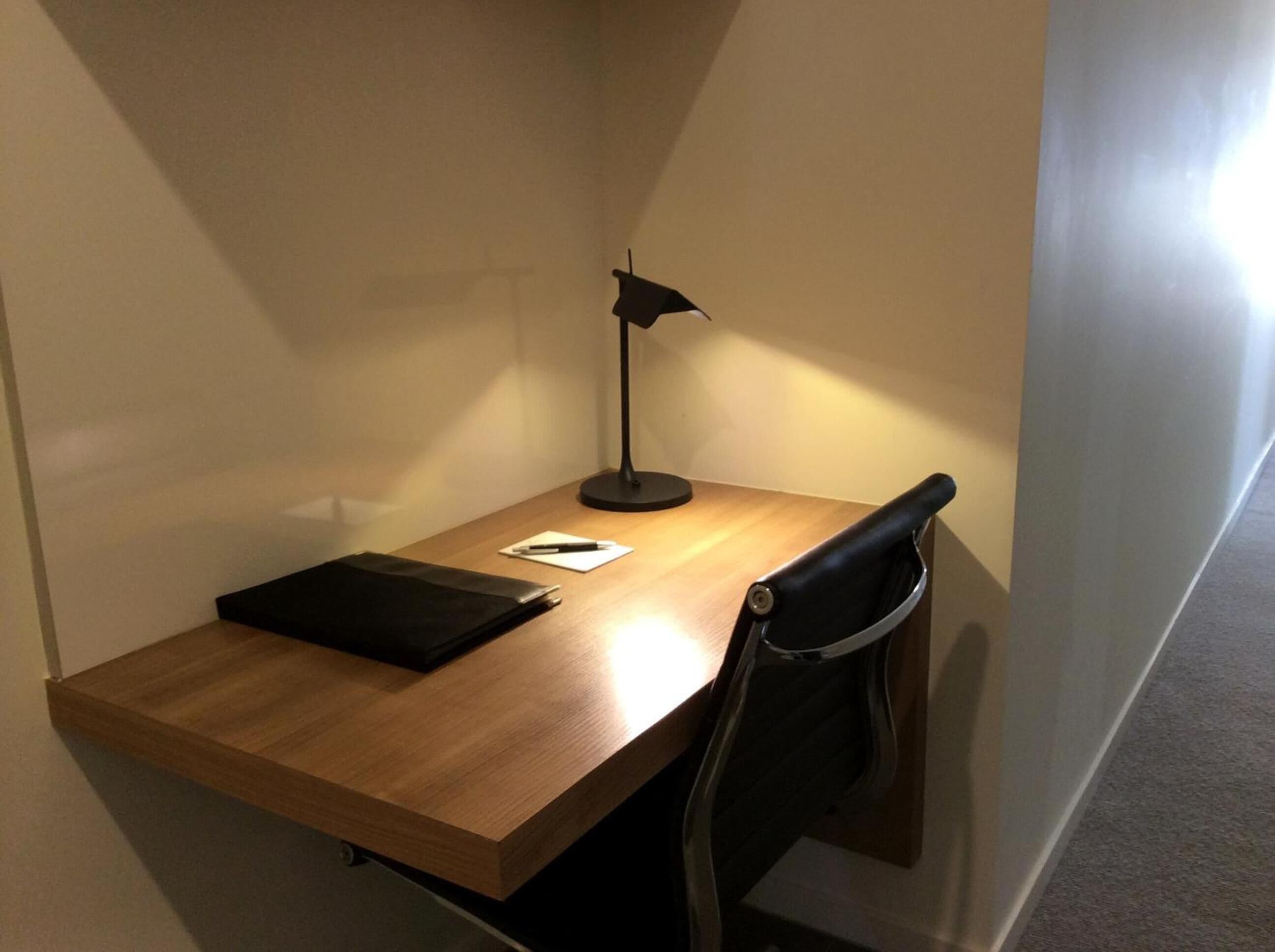 A corner desk with a black chair, a black desk lamp, and a black folder on the desk.