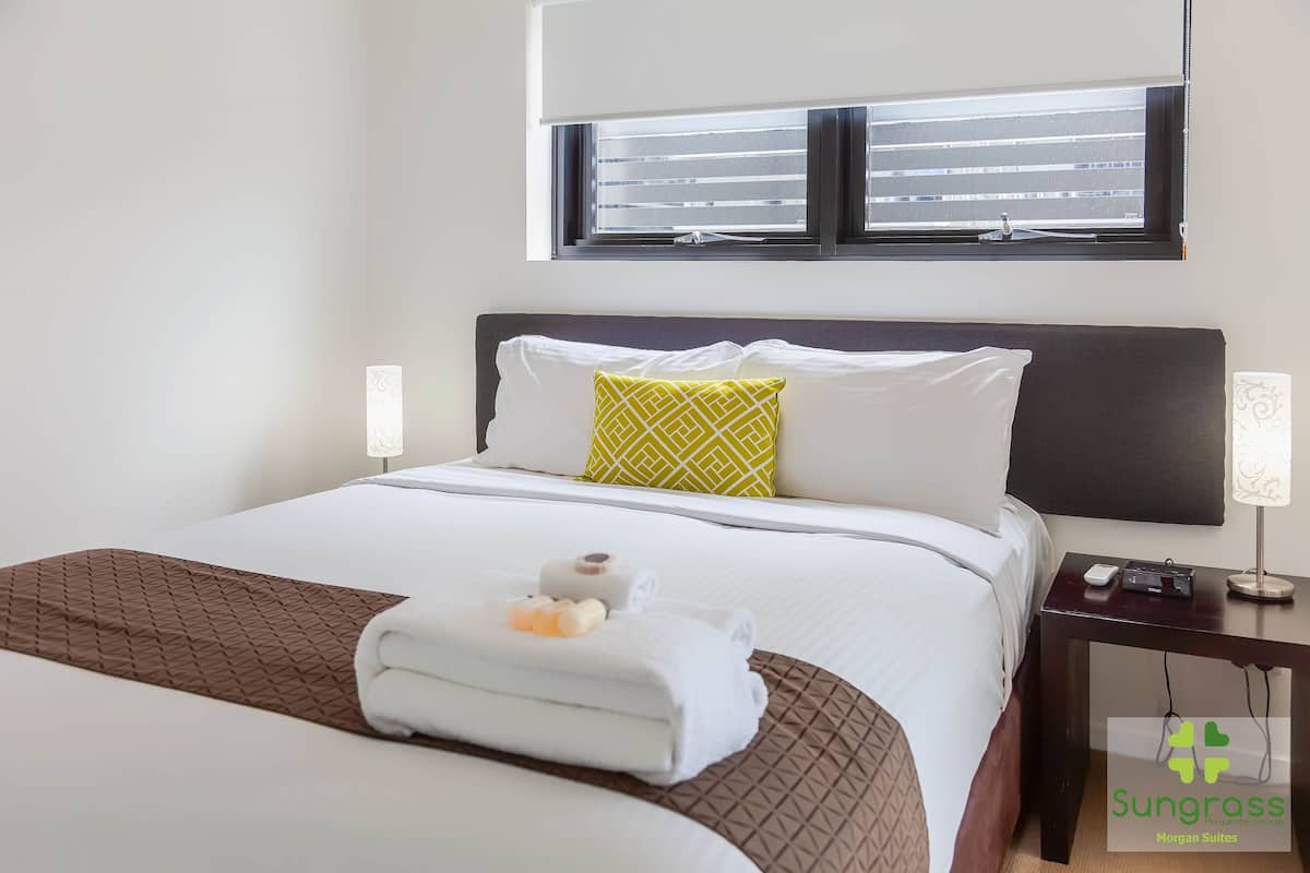 A hotel room with a bed, a nightstand, a lamp, and a window with blinds.