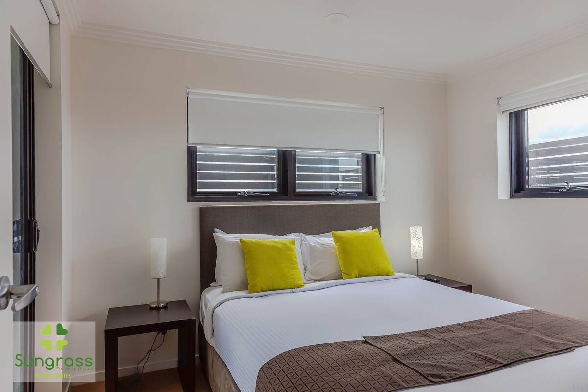 A hotel room with a bed, a bedside table, a lamp, and a window with blinds.