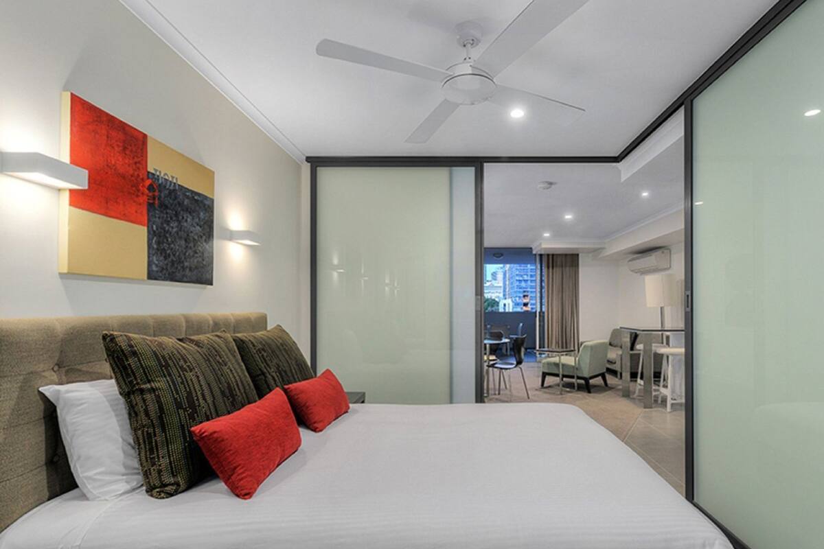 A modern hotel room with a large bed, a ceiling fan, a glass partition, and a city view.