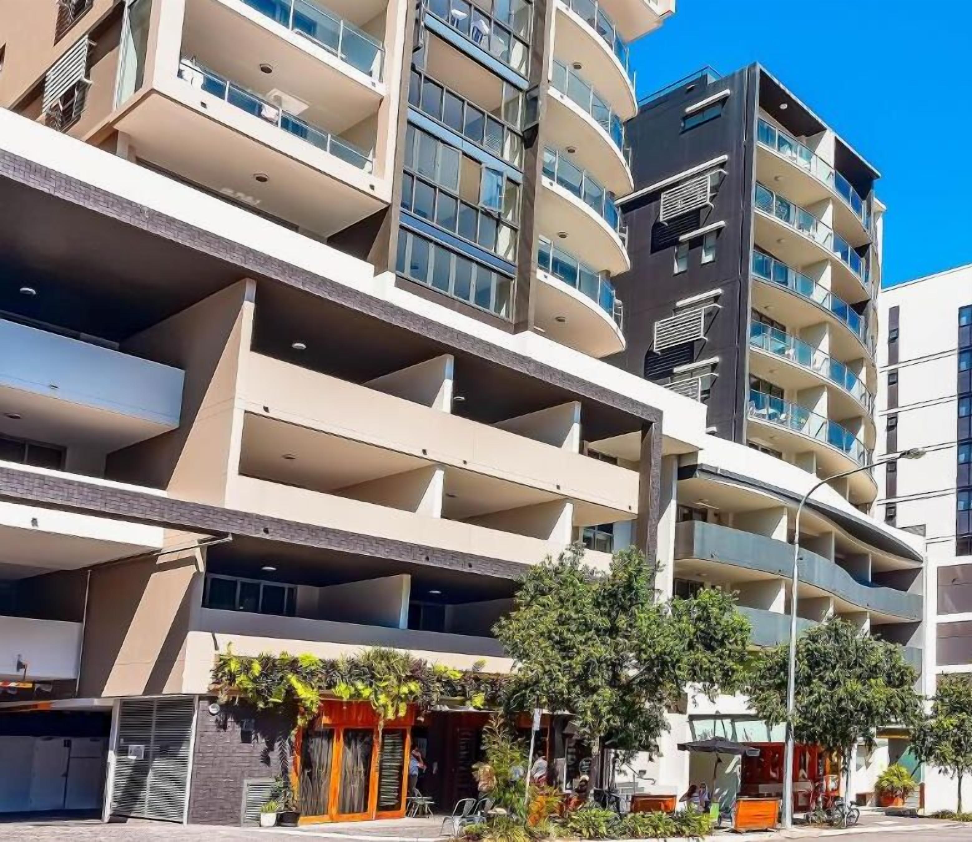 A multi-story residential building with balconies, a covered parking area, and a small outdoor seating area with plants.