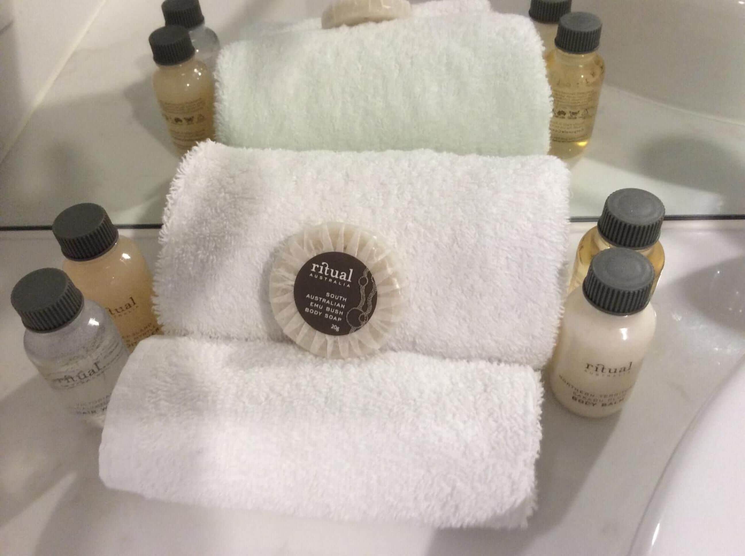 Un baño con una toalla blanca, un albornoz y varias botellas de Jabón Corporal Ritual Australian dispuestas sobre una superficie de vidrio.