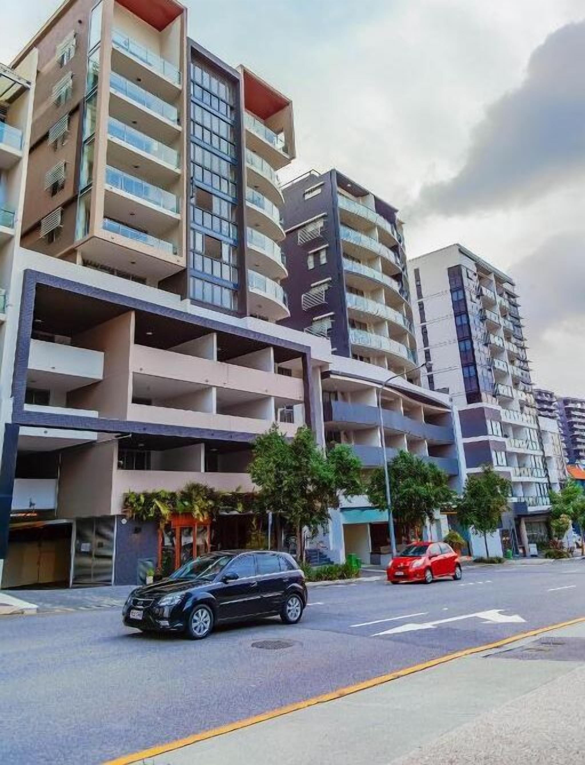 A multi-story residential building with balconies, a parking area, and a street view.