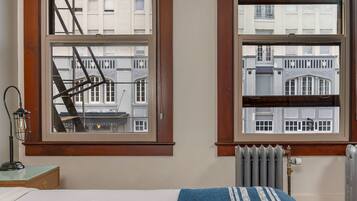 A room with a bed, a radiator, a window with a view of buildings, and a lamp on a side table.
