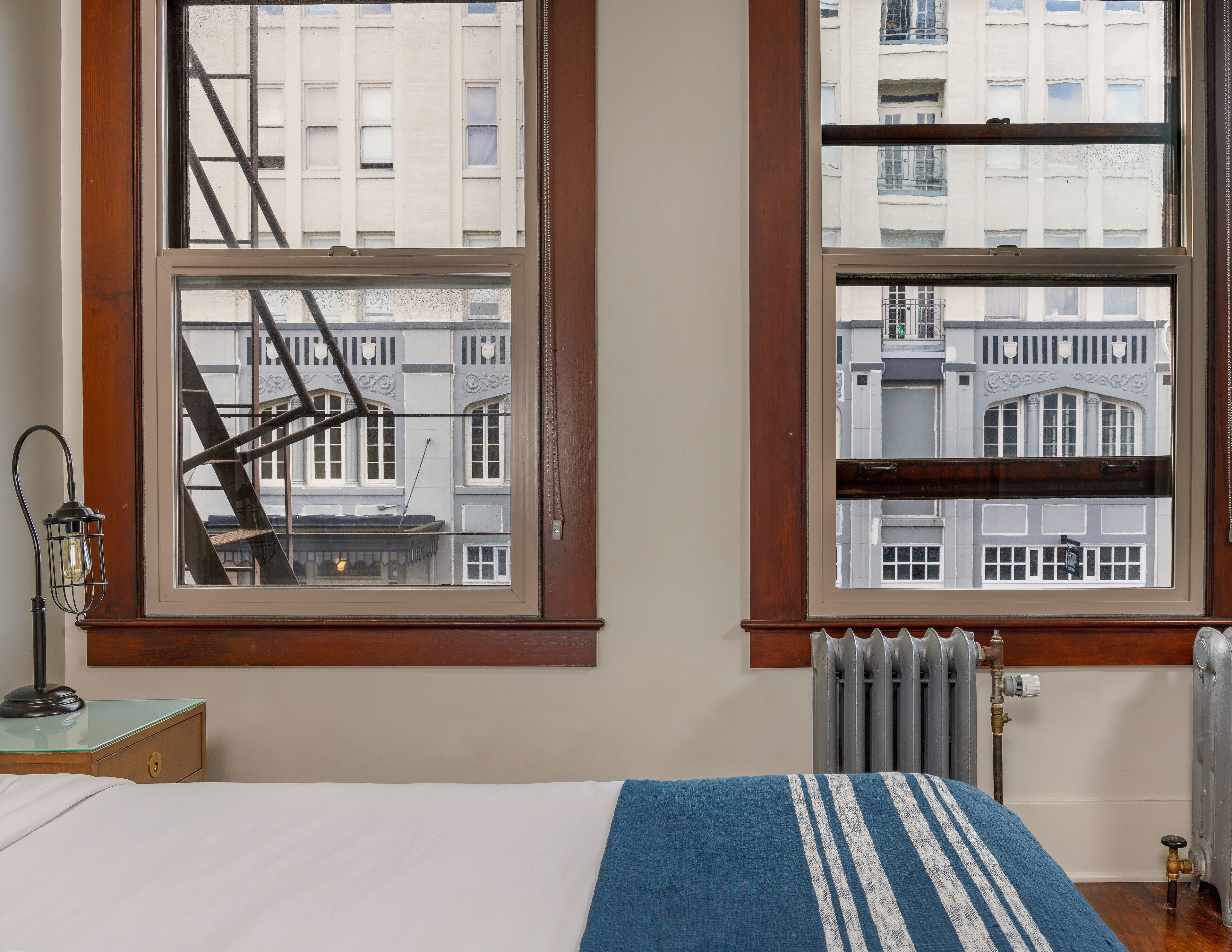 A bedroom with a bed, a nightstand, a lamp, and a view of a building through two windows.