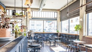 A restaurant interior with a bar area, multiple tables, chairs, hanging lights, and large windows. - Commodore Hotel (Astoria)