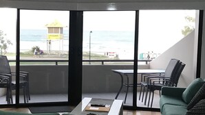 Et værelse med en grøn sofa, træstole, et bord og en ventilator i loftet. Store vinduer giver udsigt til stranden og en redningsplatform.
