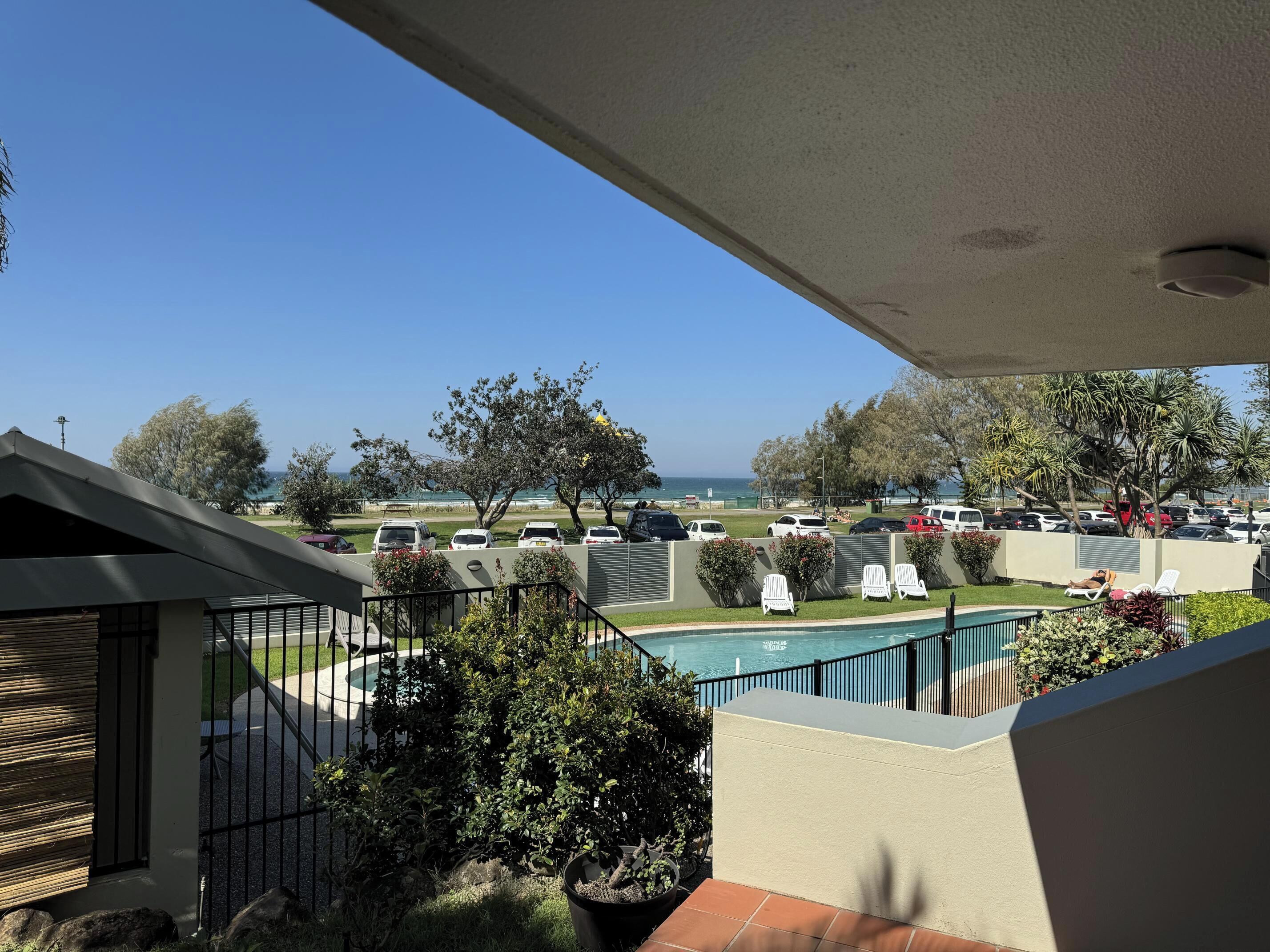 A balcony with a view of a swimming pool, parked cars, and a beach.