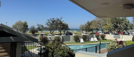 A balcony with a view of a swimming pool, parked cars, and a beach.
