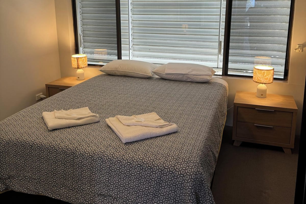 A neatly made bed with a patterned bedspread, two bedside lamps, and a window with blinds.