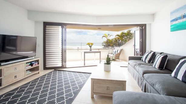 En moderne stue med fjernsyn, sofa, sofabord og udsigt til stranden gennem store skydedøre.