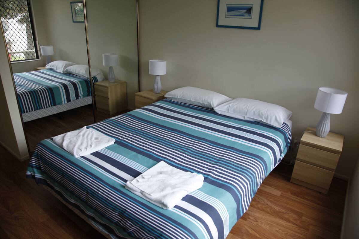 A hotel room with two beds, each with striped bedding and white pillows. There are bedside tables with lamps and a small drawer.