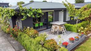 Une maison moderne dotée d’un patio comprenant une table et des chaises en bois, le tout entouré d’une végétation luxuriante et de plantes en fleurs.