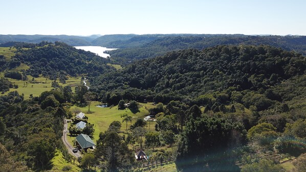Un paysage rural avec un lac, une forêt dense et quelques bâtiments, dont une maison avec un toit vert et une petite construction surmontée d’un drapeau.