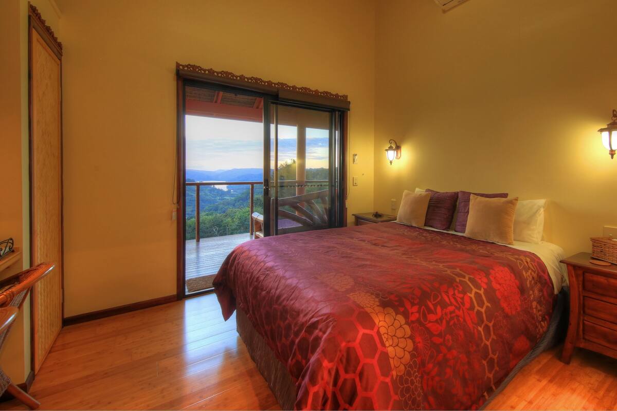 A bedroom with a large bed, a wooden nightstand, a vase with flowers, and a view of the outdoors through a sliding door.